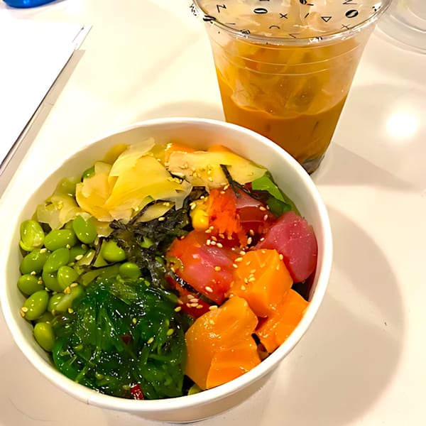 A vibrant poke bowl with tuna, salmon, edamame, seaweed salad, and pickled ginger, next to a drink.